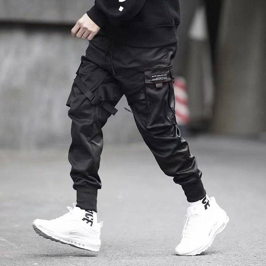 Side view of men's black cargo jogger pants with functional pockets and ribbon straps, paired with white sneakers for casual style.