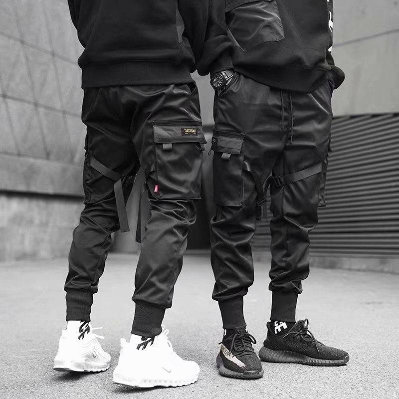 Urban streetwear men's cargo jogger pants in black with multi-pocket design and strap details, styled with black and white sneakers.