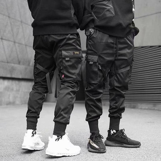 Urban streetwear men's cargo jogger pants in black with multi-pocket design and strap details, styled with black and white sneakers.