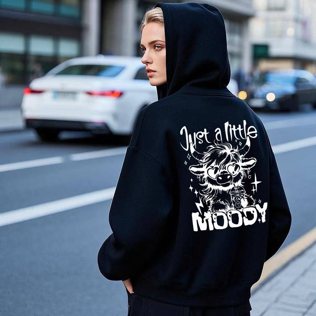 Black American men's and women's casual trend fashion printed hoodie with 'Just a Little Moody' cartoon cow design, worn outdoors on city street.