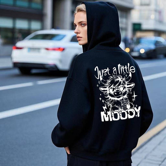Black American men's and women's casual trend fashion printed hoodie with 'Just a Little Moody' cartoon cow design, worn outdoors on city street.